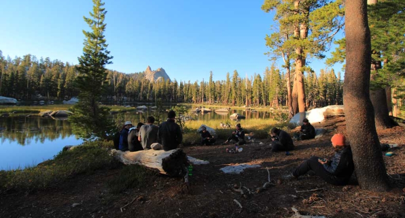 A group of students sit in a wooded area near a lake. In the distances, there is a mountain. 